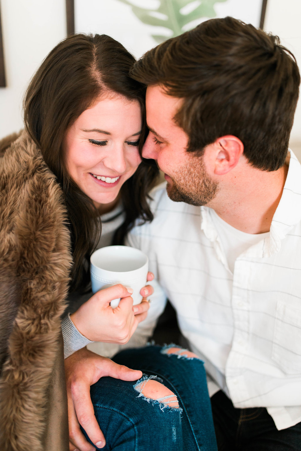 a newly engaged couple sitting on a couch while sipping coffee, snuggling, and laughing