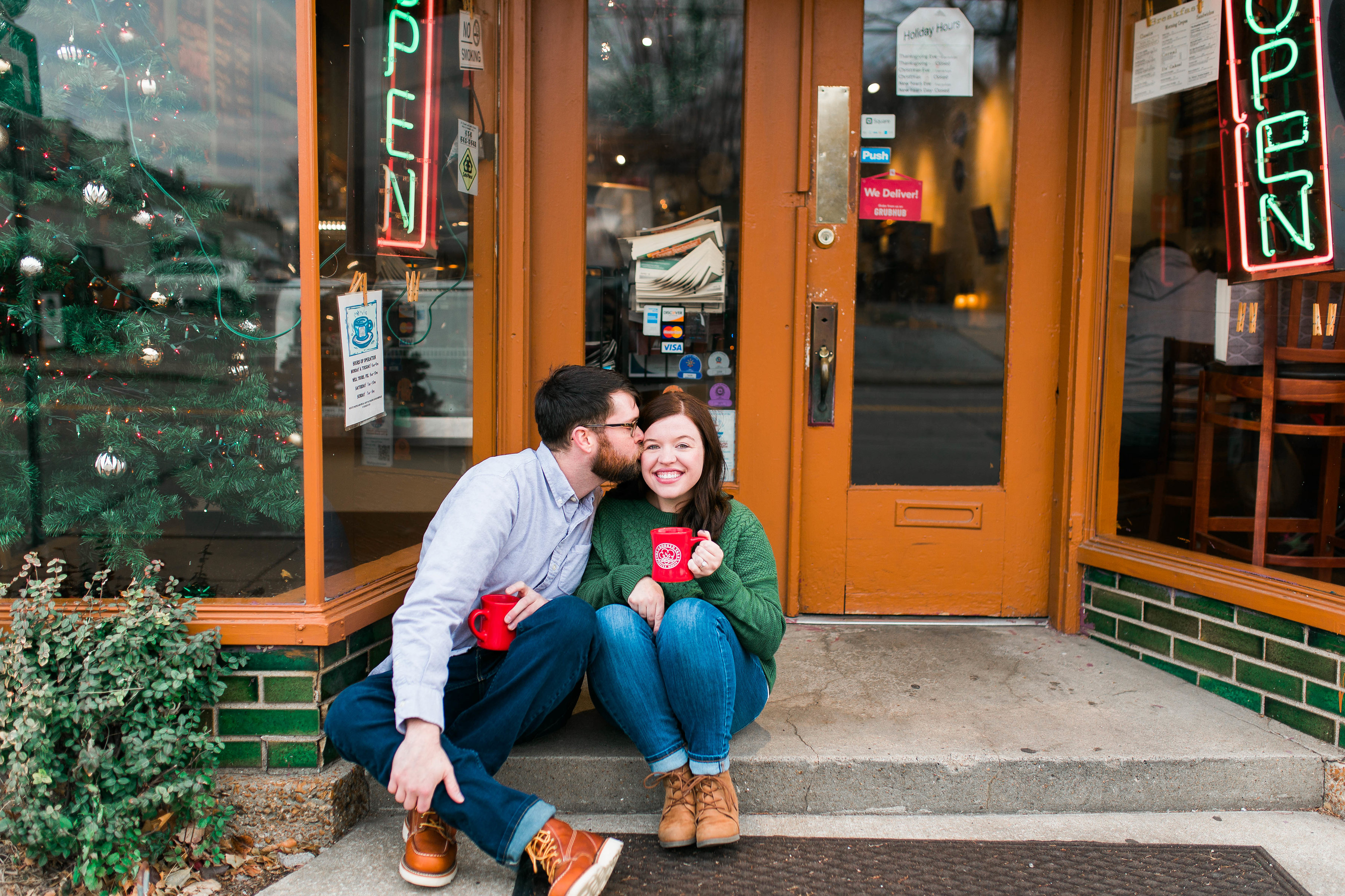 a newly engaged couple drinking out of red coffee mugs on the step outside of a coffee shop