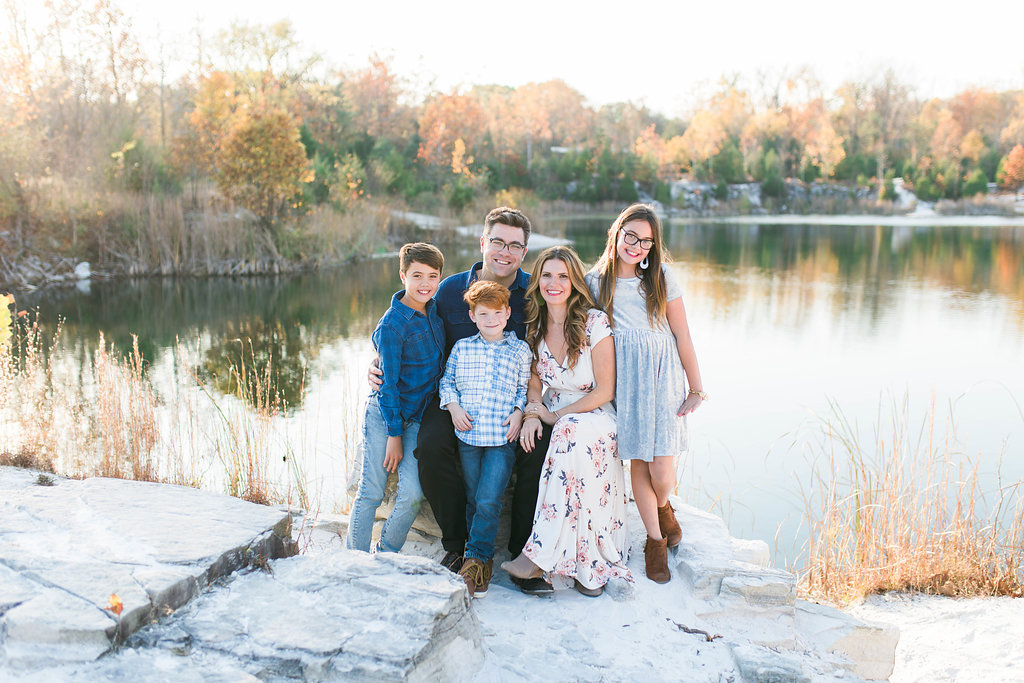 a family of five, one girl and two boys, smiling together infront of a pretty lake