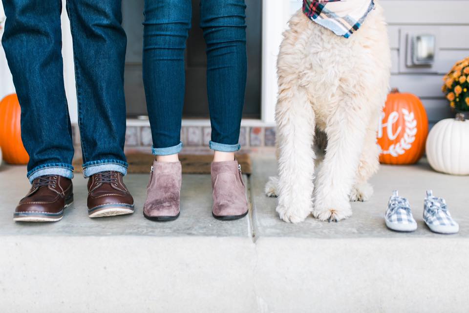 the legs of a soon to be mom, dad, their soft coated wheaten terrior, and plaid blue baby shoes