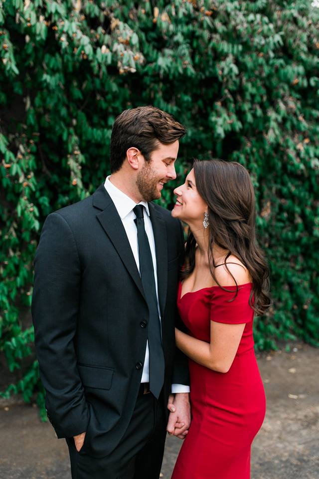 newly engaged woman in a red dress and man in suit laughing with eachother with greenery in the background