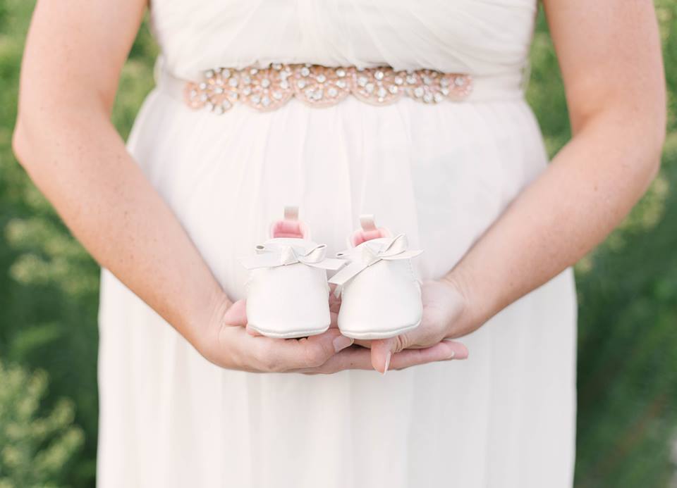 a pregant woman wearing a light pink dress, holding white baby shoes infront of her belly