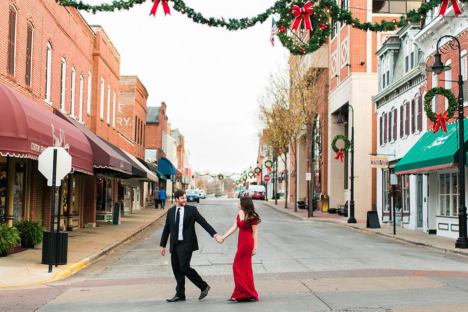a couple, a woman in a long red dress, and a man in a suit, walking across a street with Christmas decorations all around