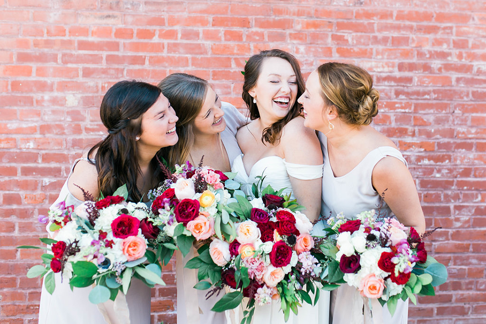 three brunette bridesmaids in cream colored dresses and a bride laughing together infront of a red brick wall, holding bouquets of red, pink, and white roses