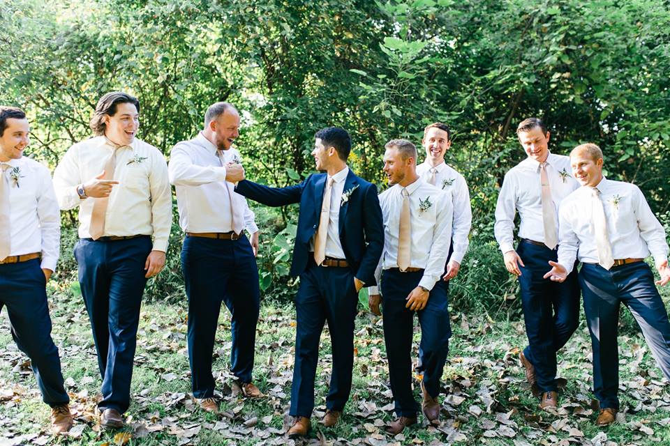 7 groomsmen and a groom laughing and messing around with greenery in the background