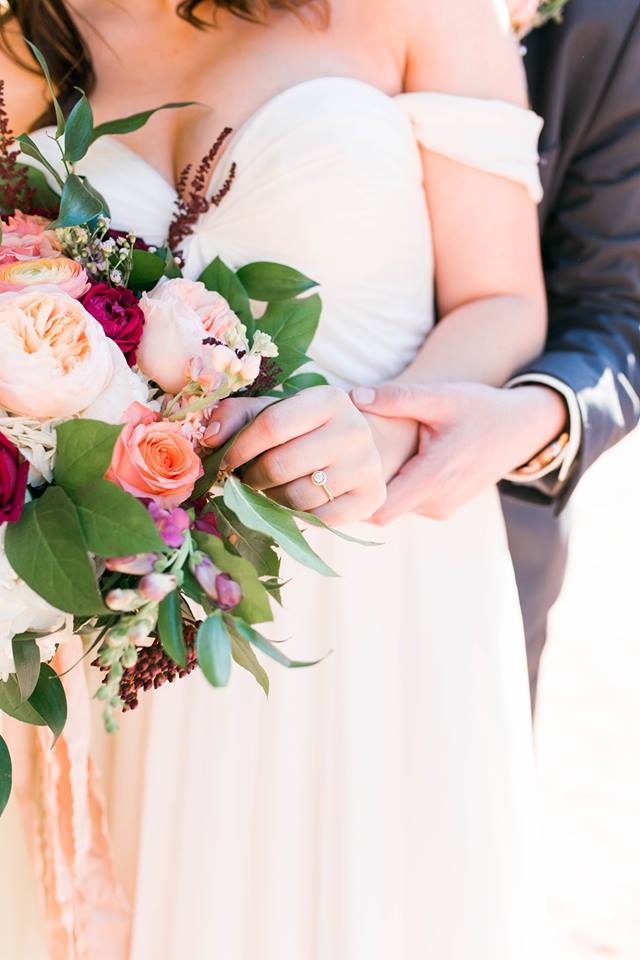 a bride holding pink and white rose bouquet with her groom behind her holding her wedding ring hand